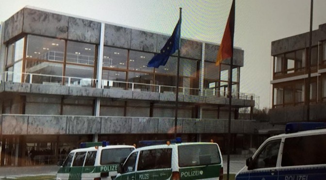 das Bundesverfassungsgericht mit Europafahne und Deutschlandfahne, davor stehen drei Polizeiwagen, es scheint trübes Wetter zu sein mit etwas Wind, im Gebäude leuchten die Deckenlampen